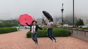 Trang (bên trái cầm ô đỏ) nhảy vui vẻ dưới mưa cùng bạn Kiesha Morris trước khách sạn Congress, thể hiện lifestyle năng động của team Trang Designs – chuyên thiết kế web chuyển đổi cao